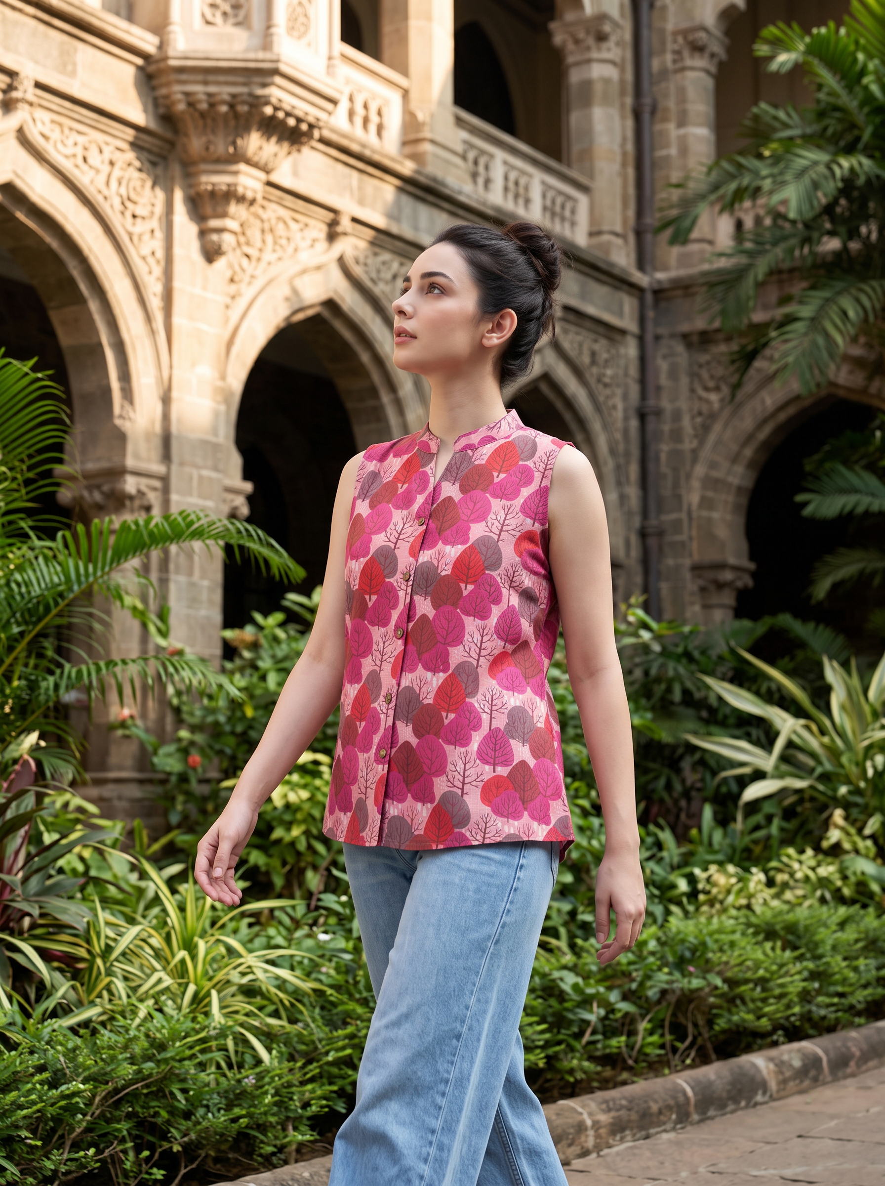 A woman wearing a sleeveless pink floral printed top and light blue jeans walks outdoors in front of a stone building with arches and lush green plants.
