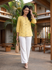 A woman walking in a modern outdoor courtyard wearing a light yellow embroidered kurta with white wide-leg trousers, holding a woven straw bag.