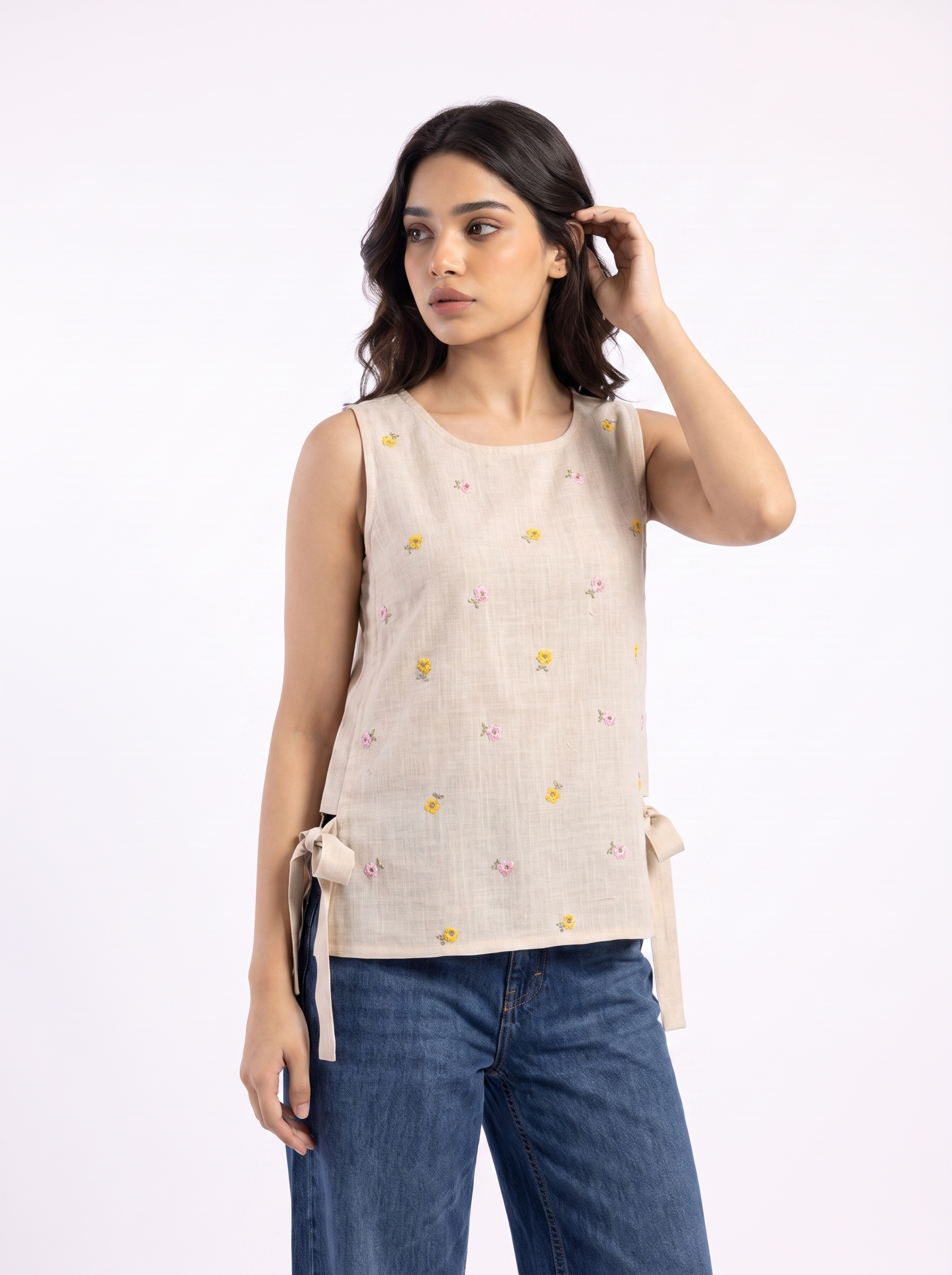 A woman modeling a sleeveless beige linen top with small yellow and pink floral prints and side tie details, paired with blue jeans, against a plain white background.