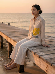 A woman sits on a wooden pier by the ocean at sunset, wearing a light beige button-up shirt with a large yellow floral print and matching wide-leg pants.