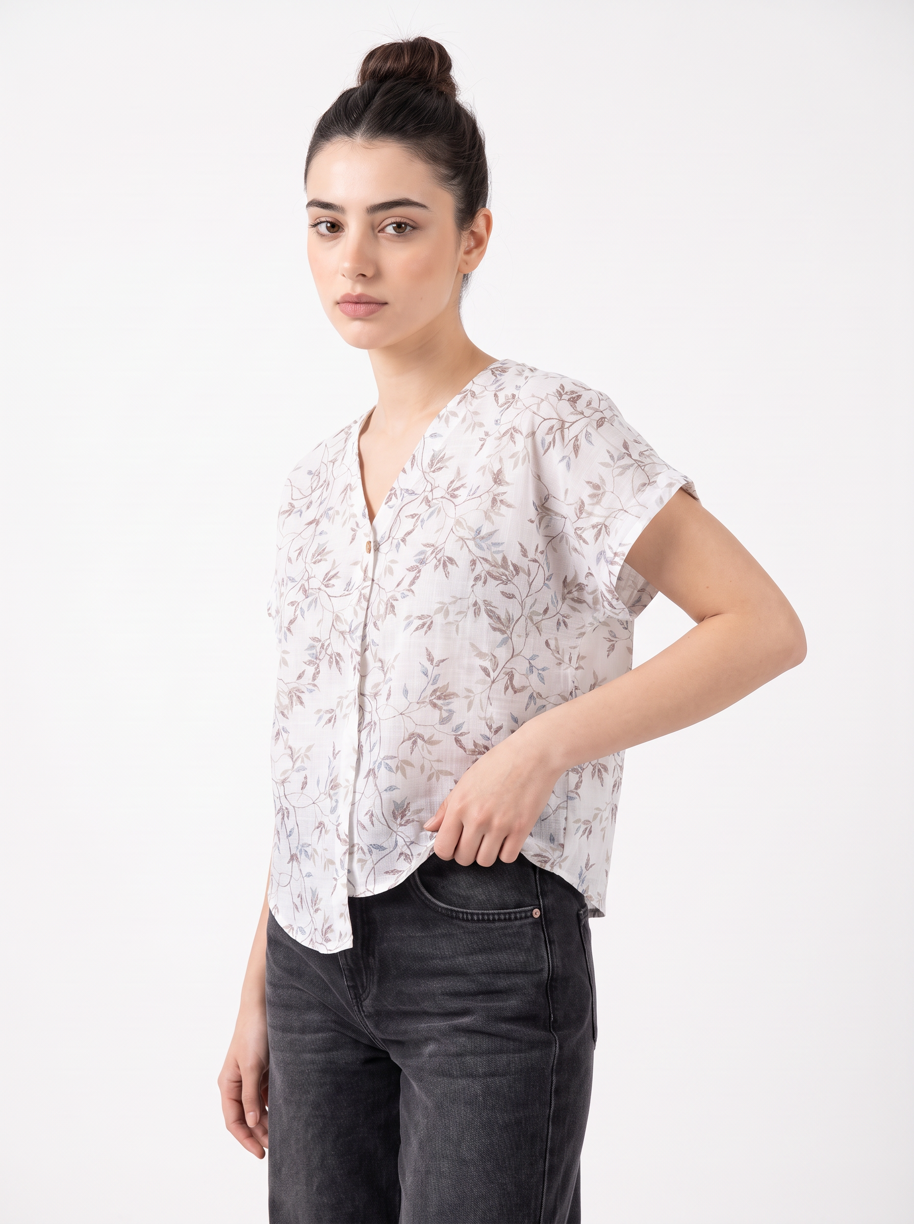 A woman modeling a short-sleeve button-up blouse with a delicate beige and brown botanical leaf pattern on a white base, paired with dark gray jeans, against a plain white studio background.