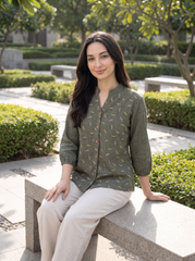 A woman sits on a stone bench in an outdoor garden setting, wearing an olive green linen button-up shirt with a yellow and white leaf pattern, paired with light beige trousers.