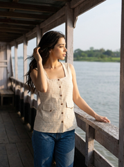 A woman stands on a wooden dock by the water wearing a sleeveless beige and white checkered top with blue jeans, looking out towards the water.