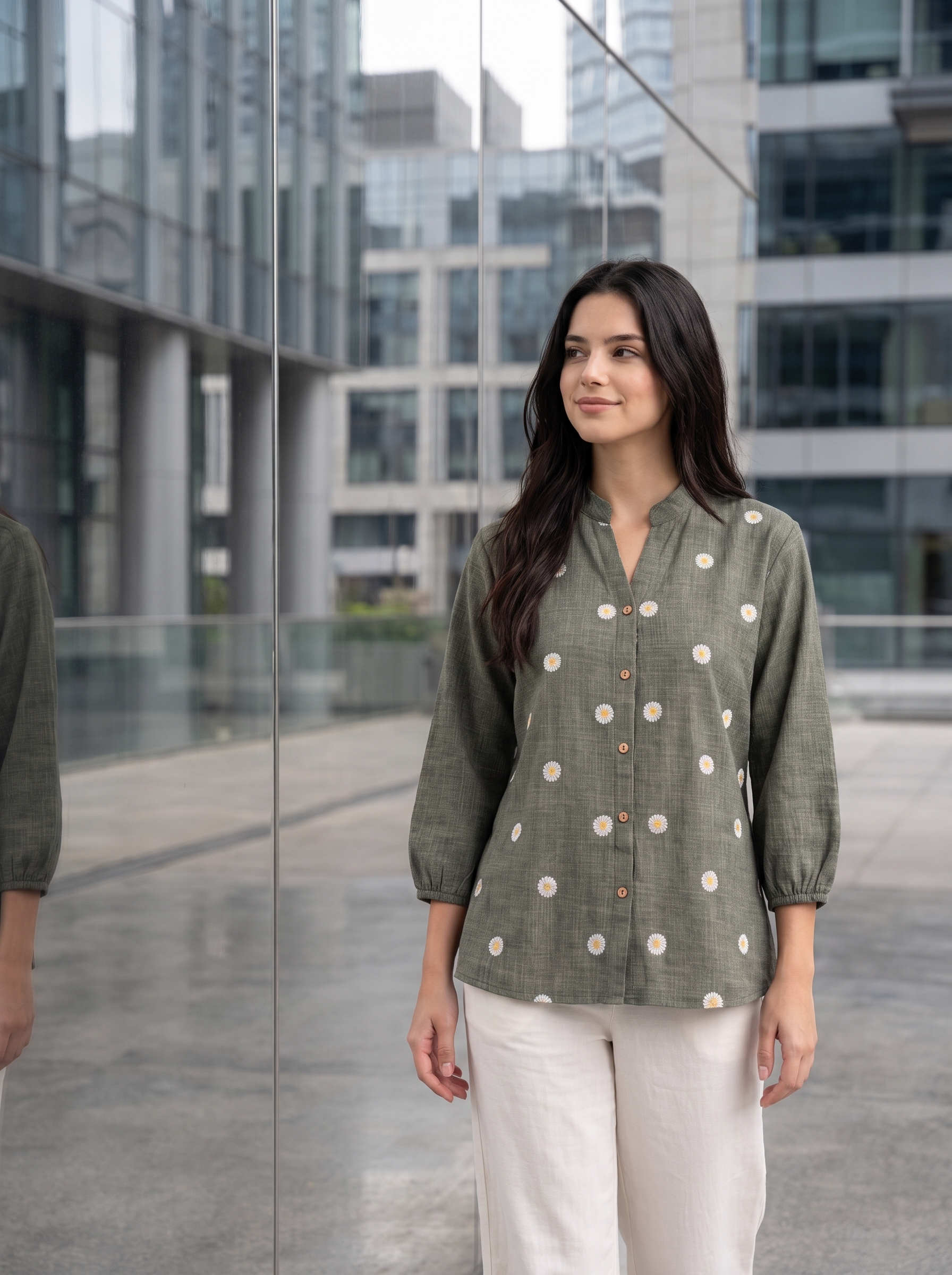 A woman wearing a sage green linen button-up blouse with white daisy patterns and white pants, standing in front of a modern glass building.