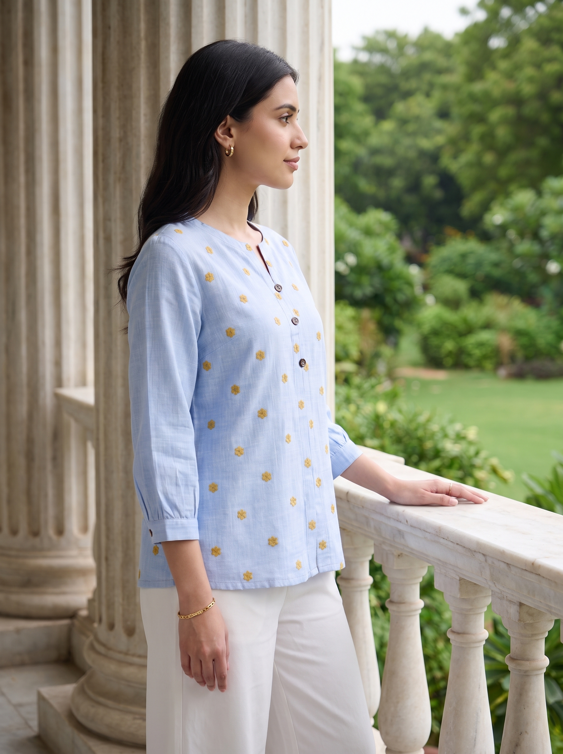 A woman stands on a balcony wearing a light blue linen kurta with small yellow floral prints and white trousers, with a garden and classical columns in the background.