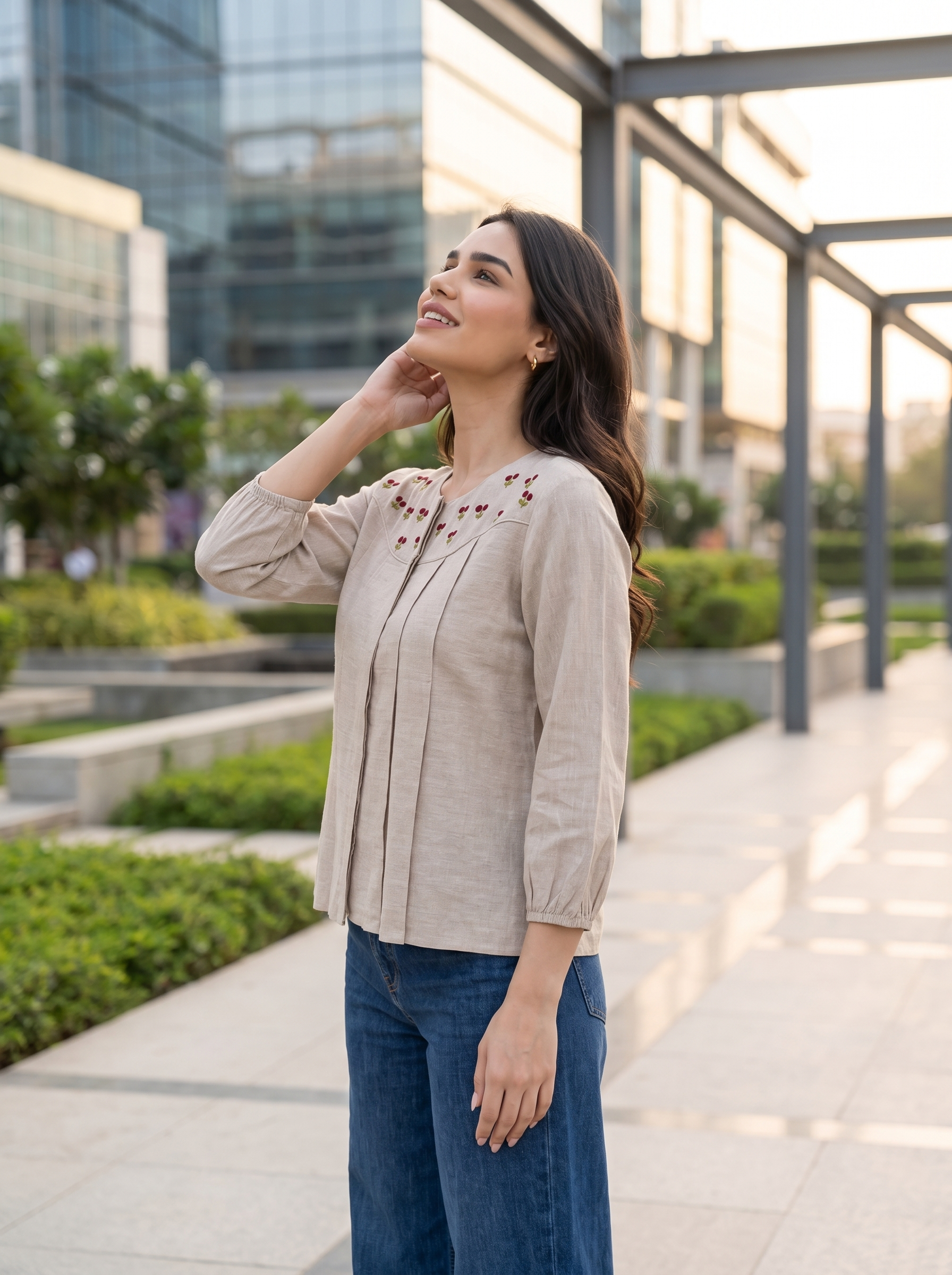 A woman wearing a beige linen blouse with brown floral embroidery on the chest and blue jeans, standing outdoors in a modern urban setting with glass buildings and a walkway.