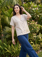 A woman wearing a light beige linen short-sleeve button-up top and blue jeans, posing outdoors in front of a lush green garden background.
