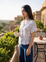 A woman wearing a white short-sleeve blouse with a delicate blue and green floral print, paired with dark blue jeans, walking along a mountain path with misty peaks in the background.