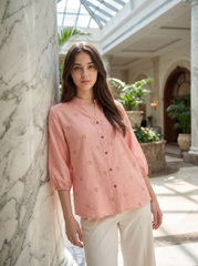 A woman stands indoors next to a large marble column, wearing a light pink button-front blouse with small embroidered motifs and cream-colored pants.