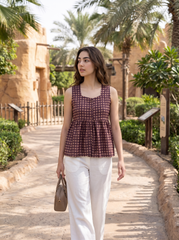 A woman stands on a sunlit pathway surrounded by palm trees and tan buildings, wearing a sleeveless burgundy and beige checkered peplum top with white pants and carrying a brown handbag.