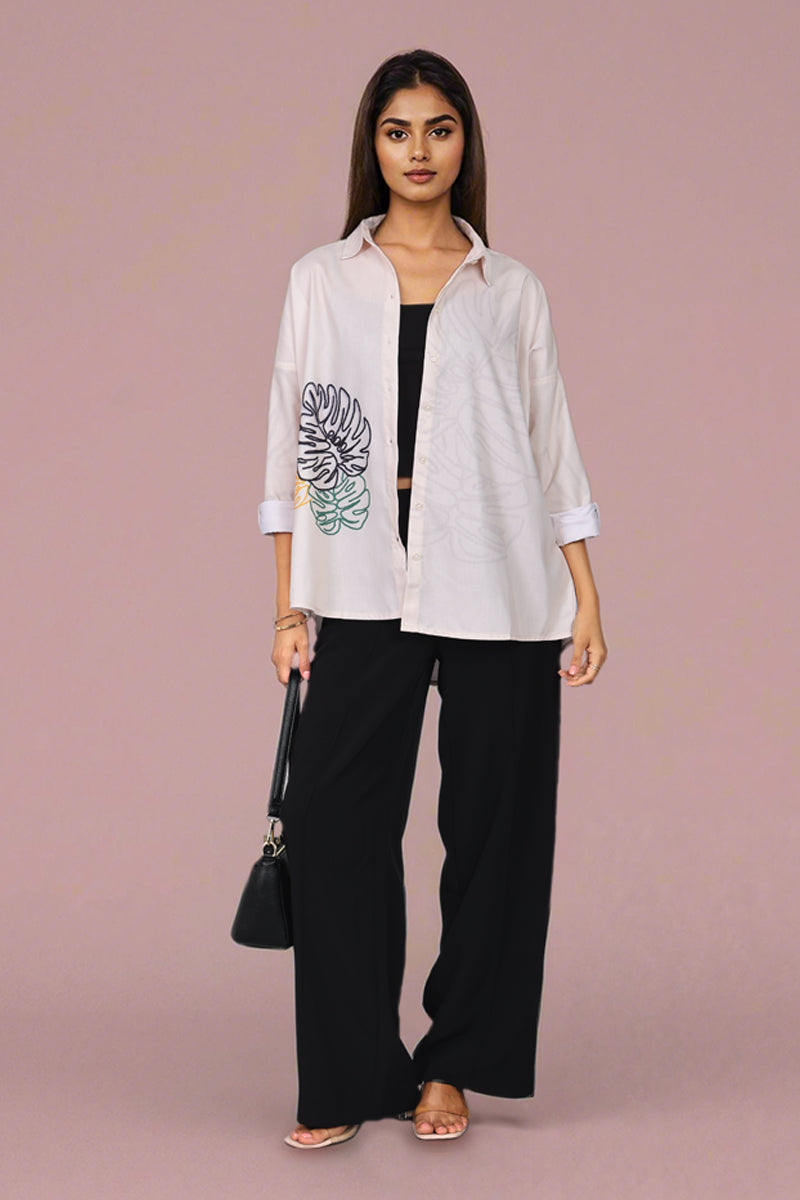 A woman models a white long-sleeve button-up shirt with a large black and green floral graphic on the left sleeve, paired with black wide-leg pants and a small black handbag, set against a solid dusty pink studio background.