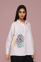 A woman modeling a white long-sleeve button-up shirt with a colorful embroidered tropical leaf design on the front, set against a solid dusty pink background.