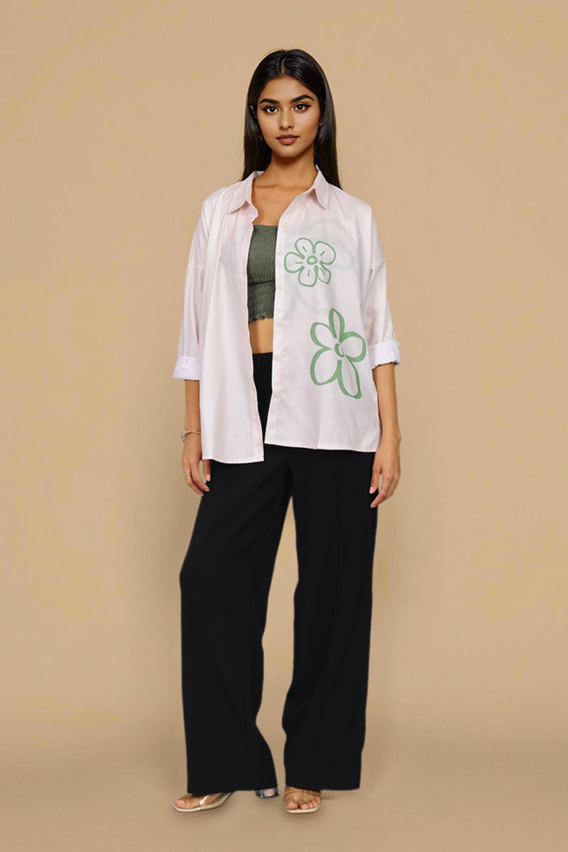 A woman stands against a plain beige studio background wearing a white button-up shirt with green floral line drawings, black wide-leg pants, a dark green crop top, and flat sandals.