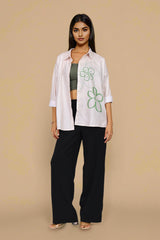 A woman stands against a plain beige studio background wearing a white button-up shirt with green floral line drawings, black wide-leg pants, a dark green crop top, and flat sandals.