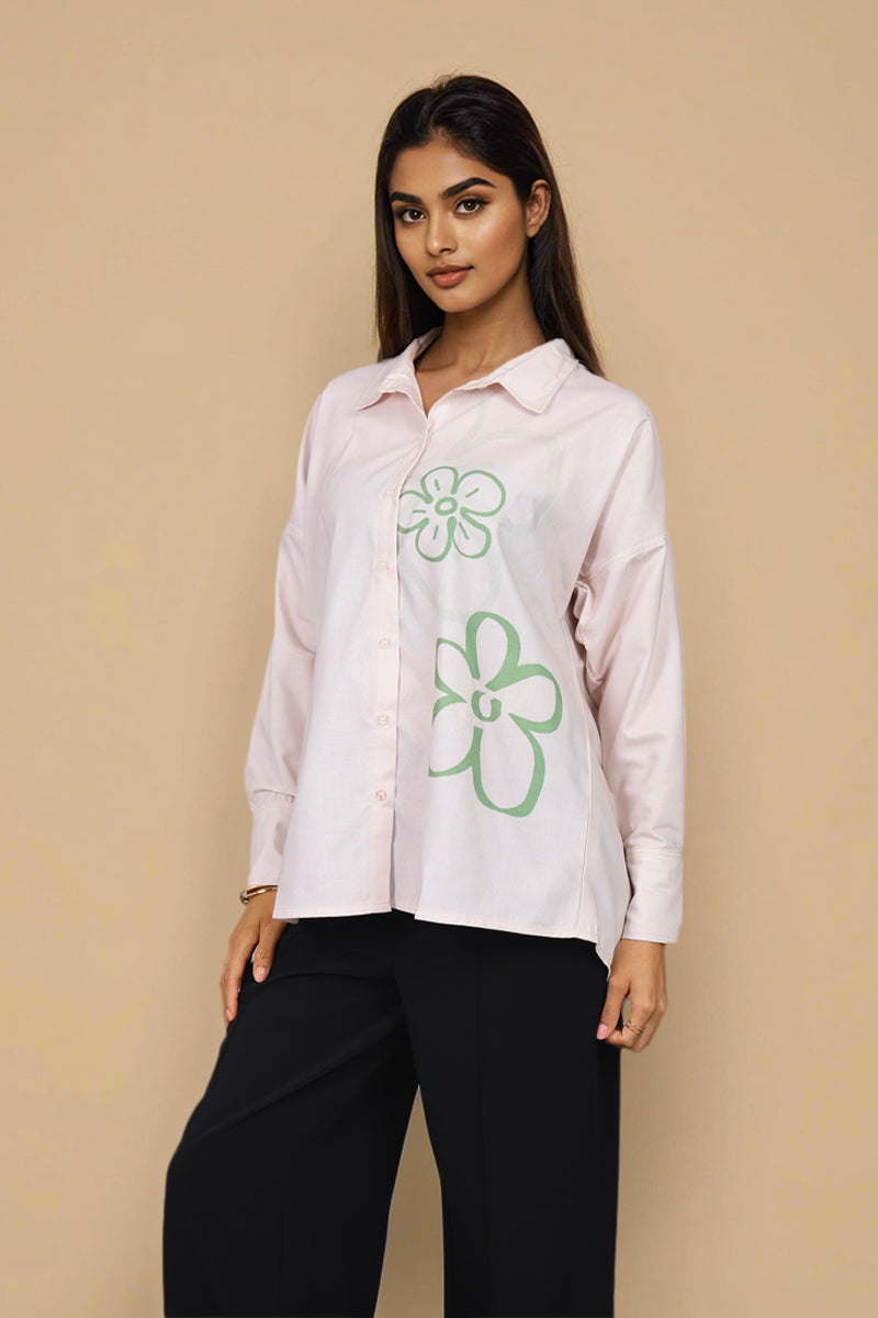 A woman modeling a light pink button-up shirt with green outline flower graphics on the front, paired with black pants, against a solid beige background.