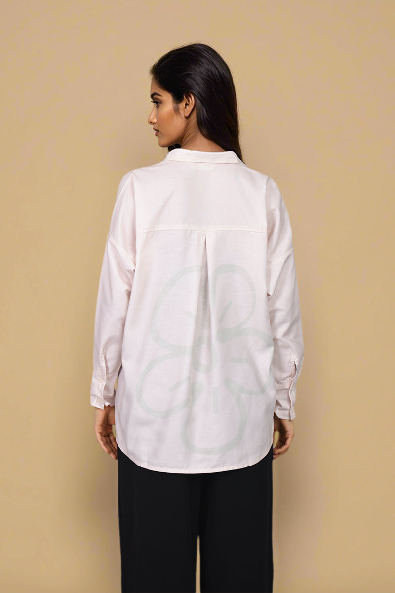 A woman shown from the back wearing a white long-sleeve button-up shirt with a large light green abstract floral pattern printed across the back, paired with black pants, against a solid beige background.