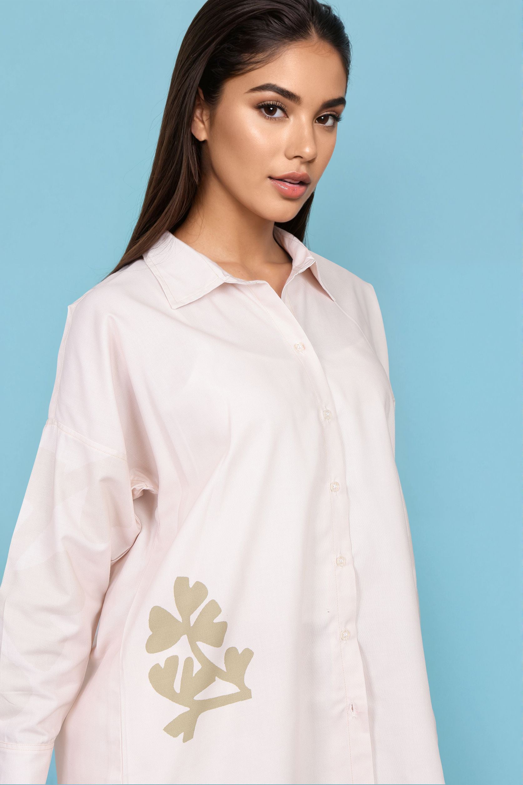 A woman wearing a light pink button-up shirt with a beige leaf embroidery on the lower left front, posed against a solid light blue background.