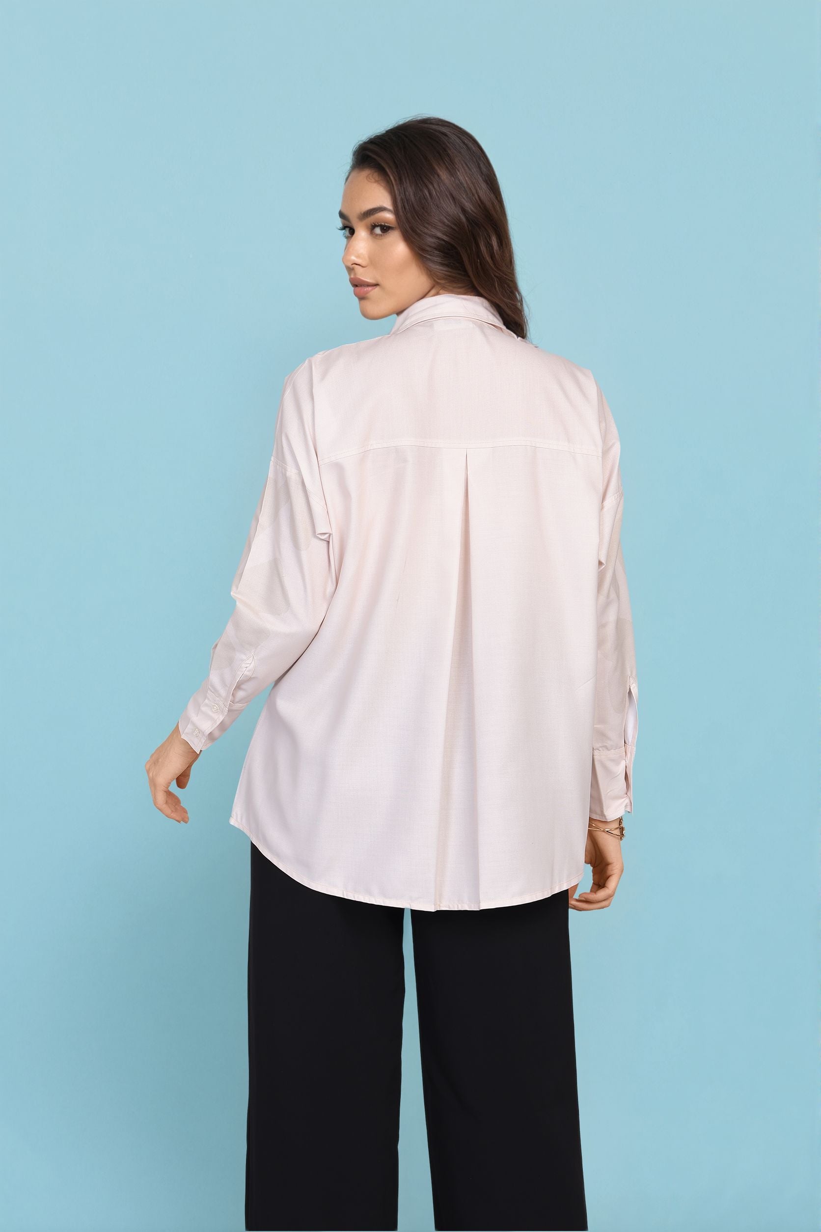 A woman shown from the back wearing a light blush pink long-sleeve button-up shirt and black wide-leg pants, standing against a solid light blue background.