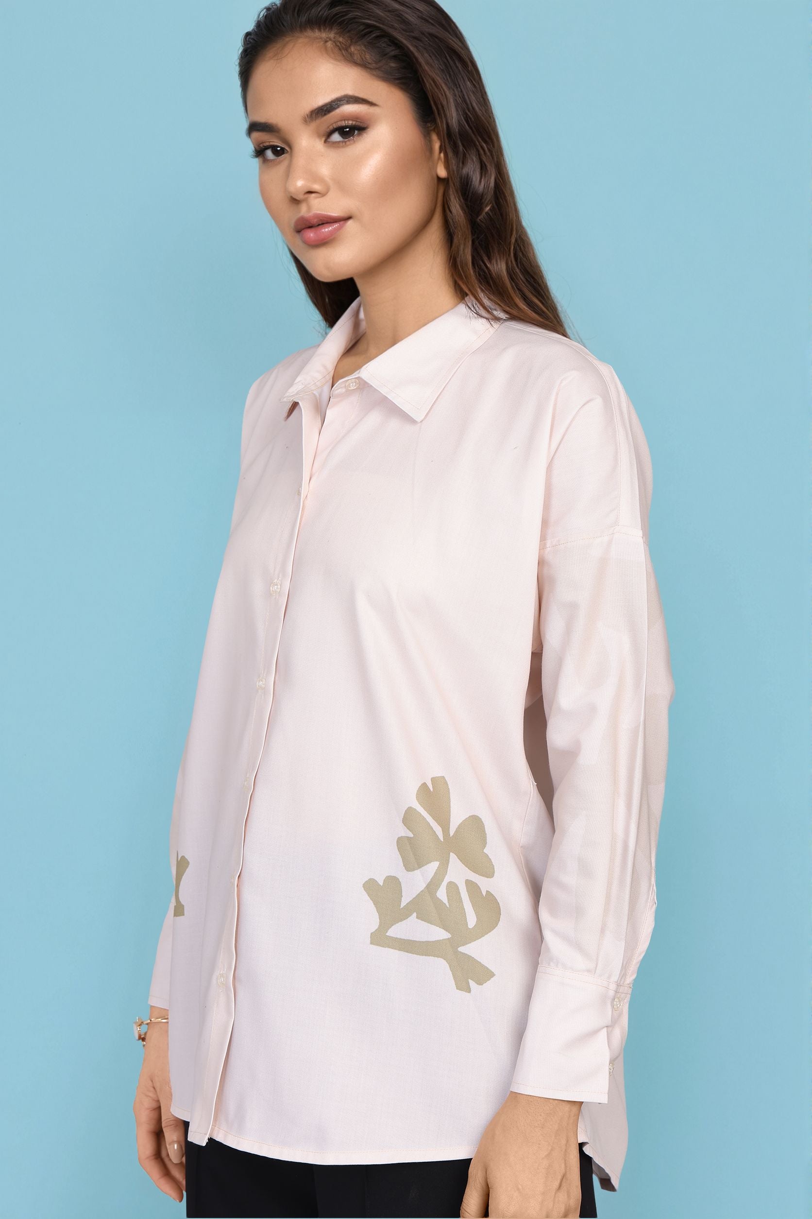 A woman wearing a light pink long-sleeve button-up shirt with gold floral embroidery on the lower front and sleeve, posed against a solid light blue background.