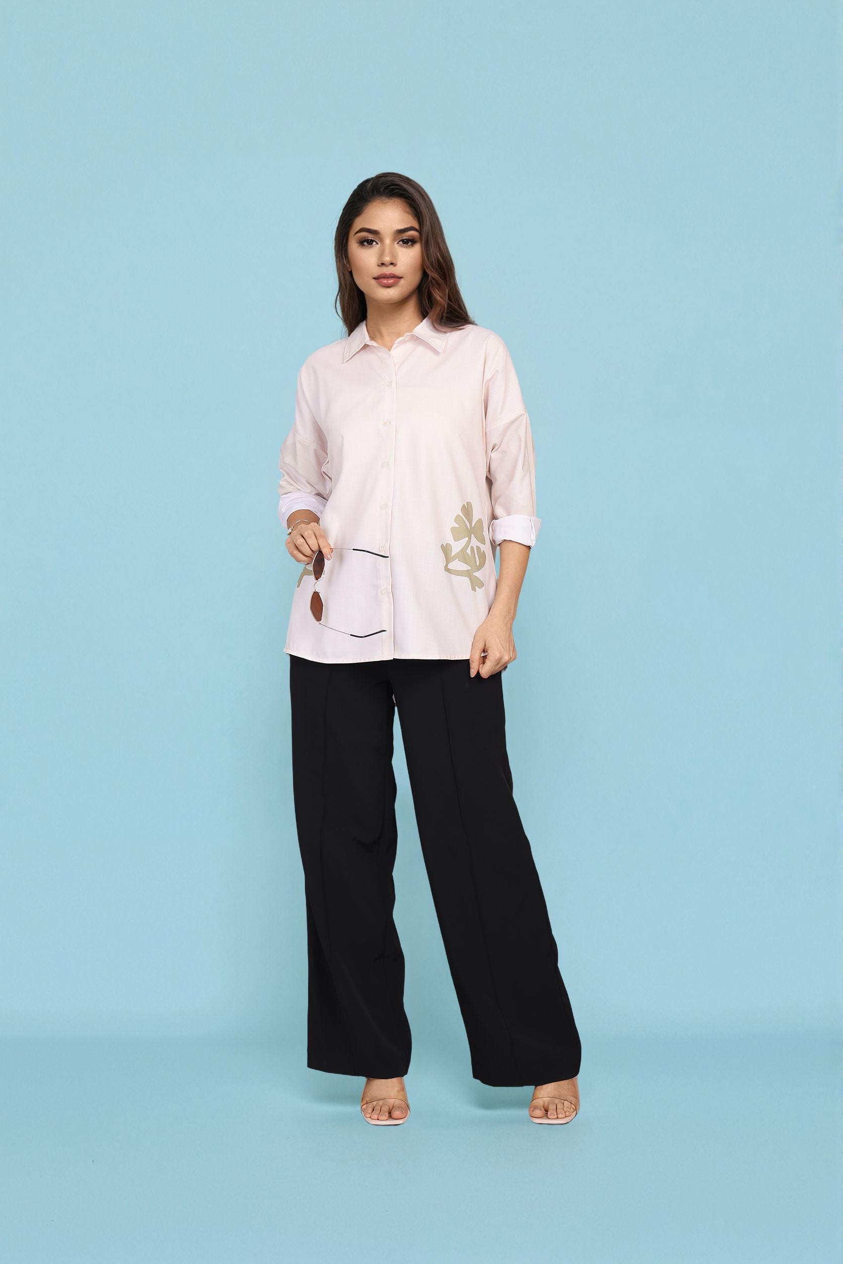A woman modeling a light pink button-up shirt with gold floral embroidery on the sleeve and front pocket, paired with black wide-leg trousers, against a solid light blue studio background.