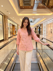 A woman wearing a light pink embroidered button-up shirt and cream trousers stands on an escalator inside a modern, upscale shopping mall.