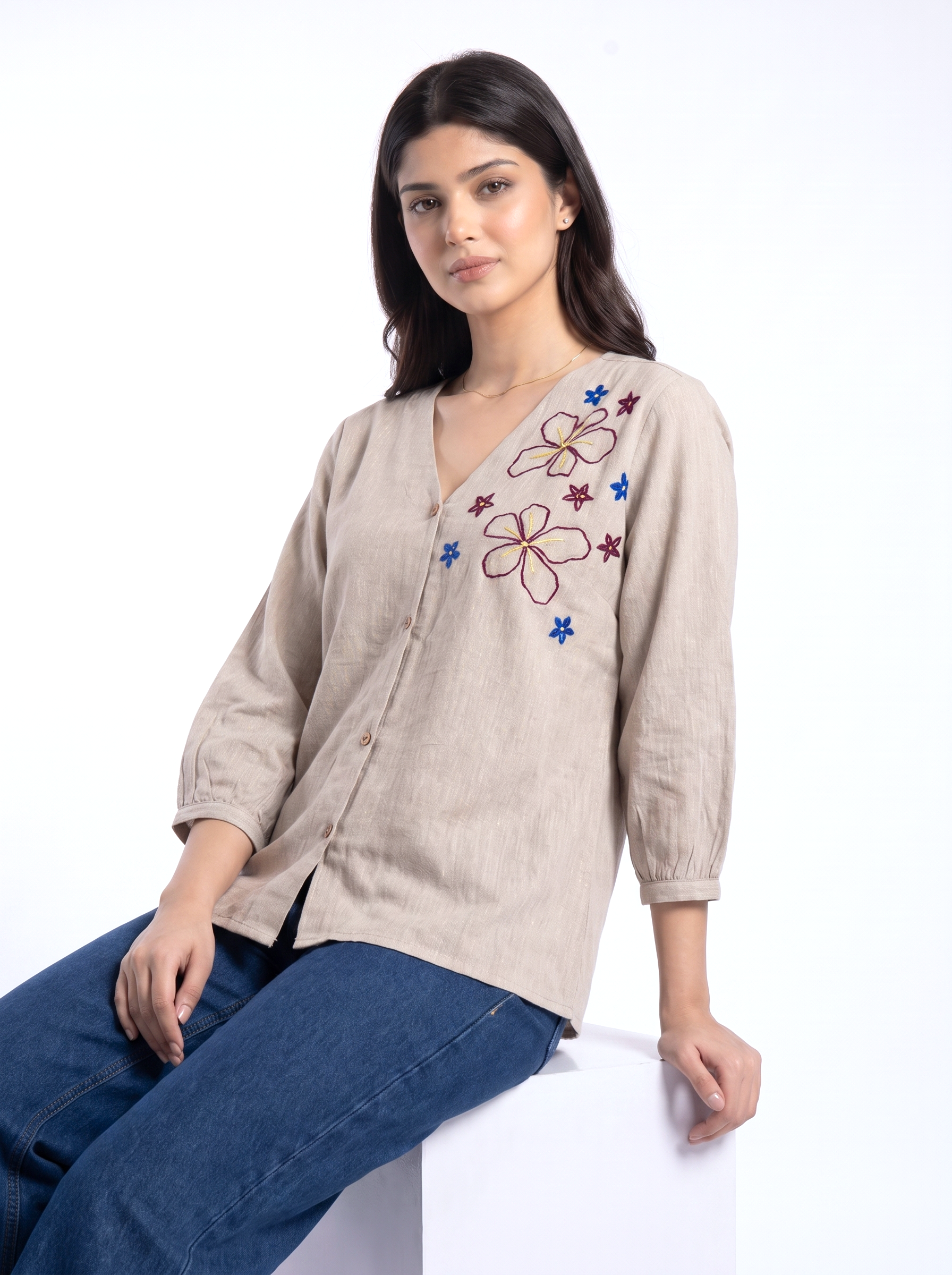 A woman wearing a beige linen button-up shirt with colorful floral and star embroidery on the chest, paired with blue jeans, seated against a plain white background.