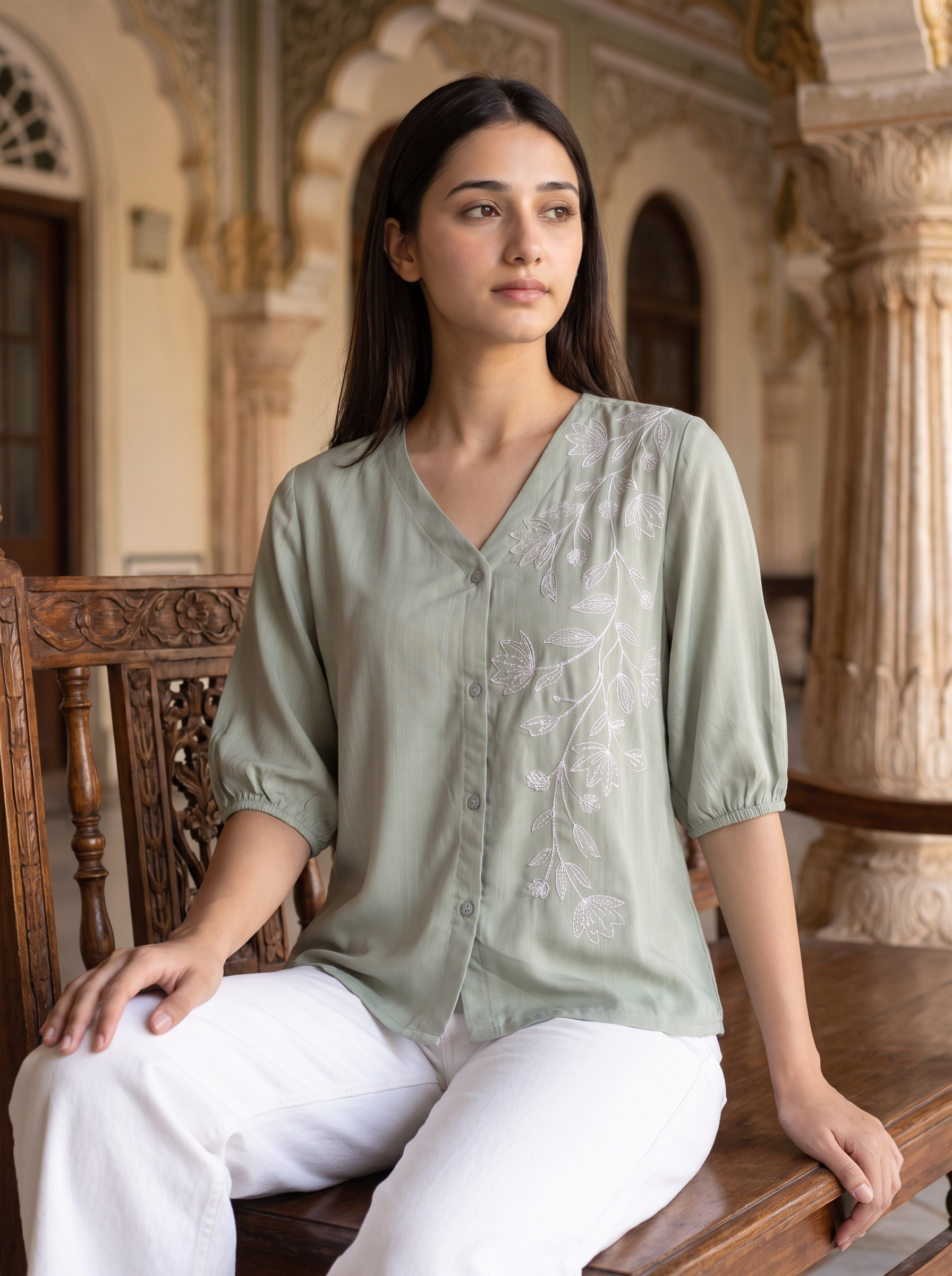 A woman sits on a carved wooden bench in an ornate indoor setting, wearing a light sage green blouse with white floral embroidery and white pants.