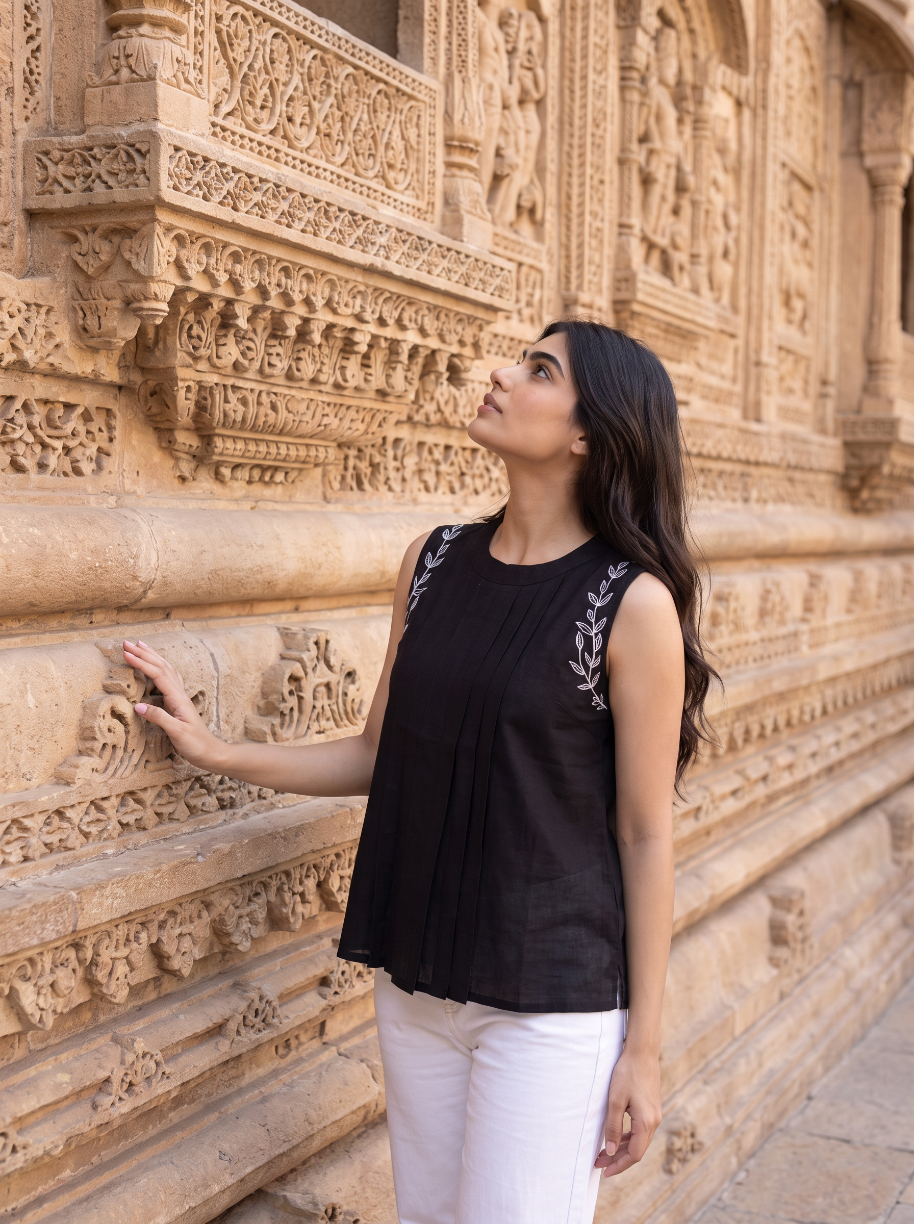 A woman wearing a sleeveless black top with white embroidery on the shoulders and white pants, standing in front of a detailed beige stone wall.