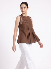 A woman modeling a sleeveless brown pleated top with white embroidered leaf details on the shoulders, paired with white pants, against a plain white studio background.