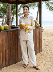A woman stands on a sandy beach under a wooden pavilion, wearing a light beige linen pajama set with a yellow lemon and green leaf graphic on the shirt. She holds a coconut with a straw.