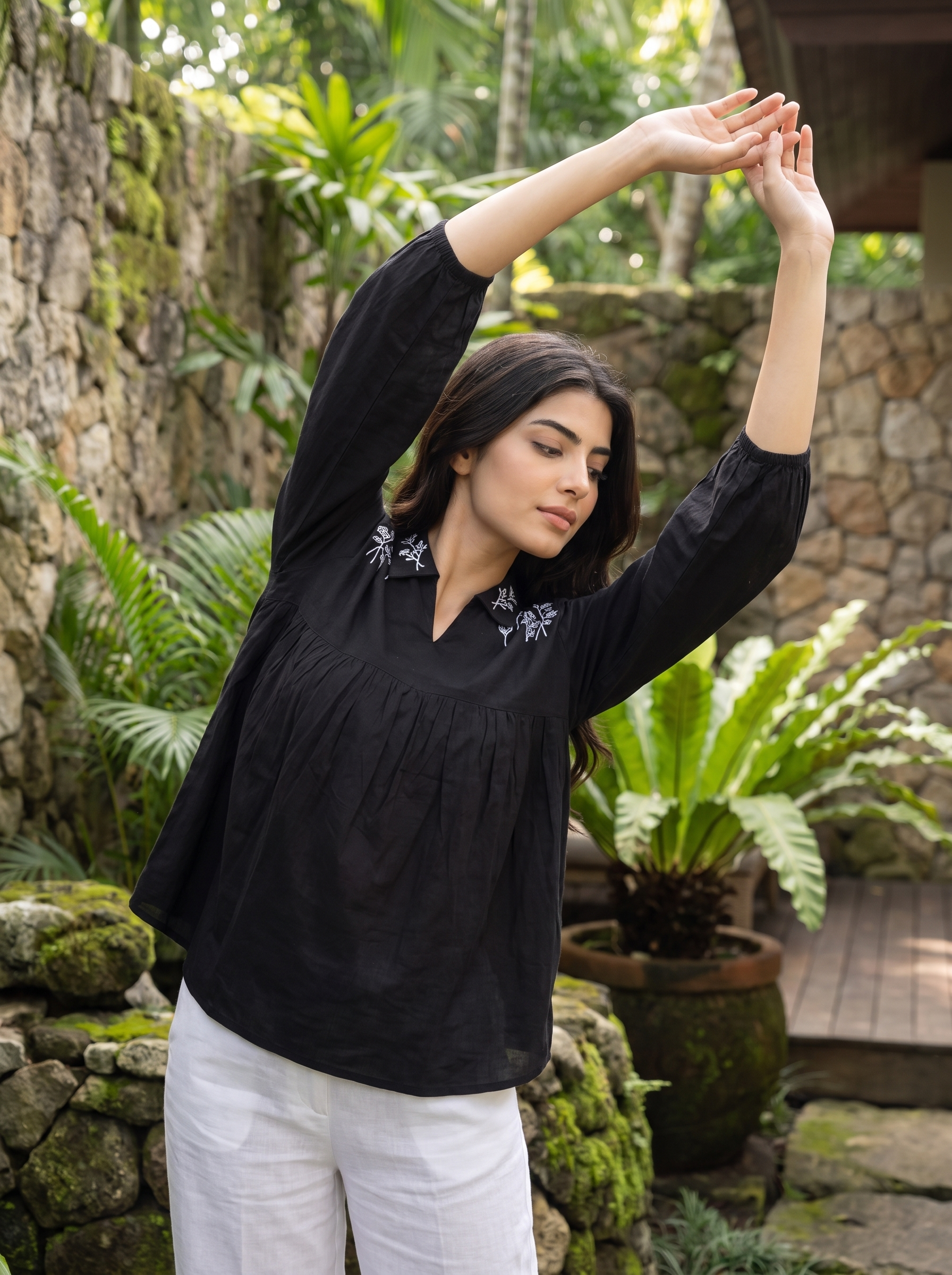A woman in a black long-sleeve blouse with white floral embroidery on the chest and white pants, stretching her arms overhead in a lush, green outdoor garden setting.