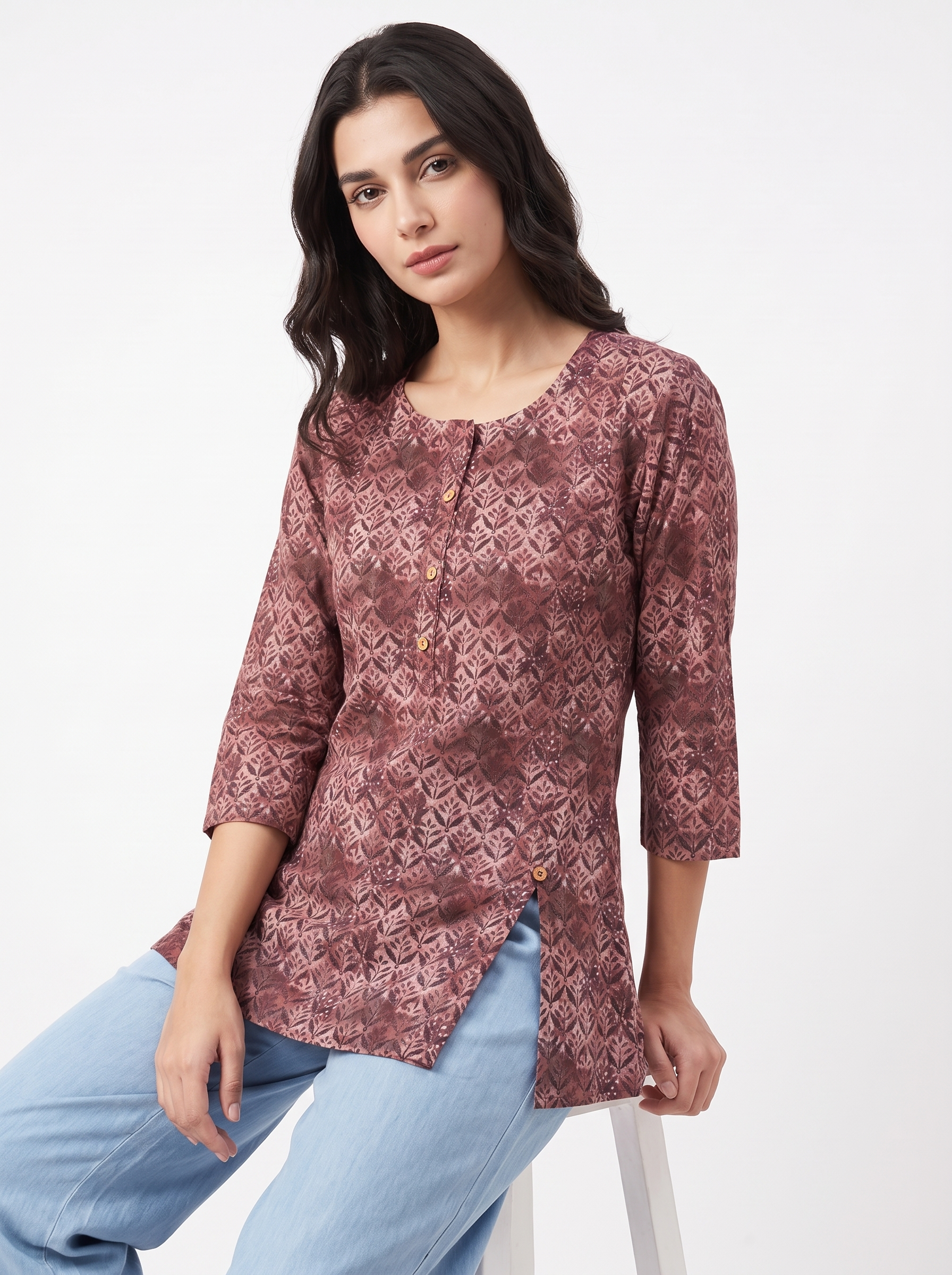 A woman wearing a dusty rose and brown patterned kurta with three-quarter sleeves, paired with light blue jeans, seated against a plain white studio background.