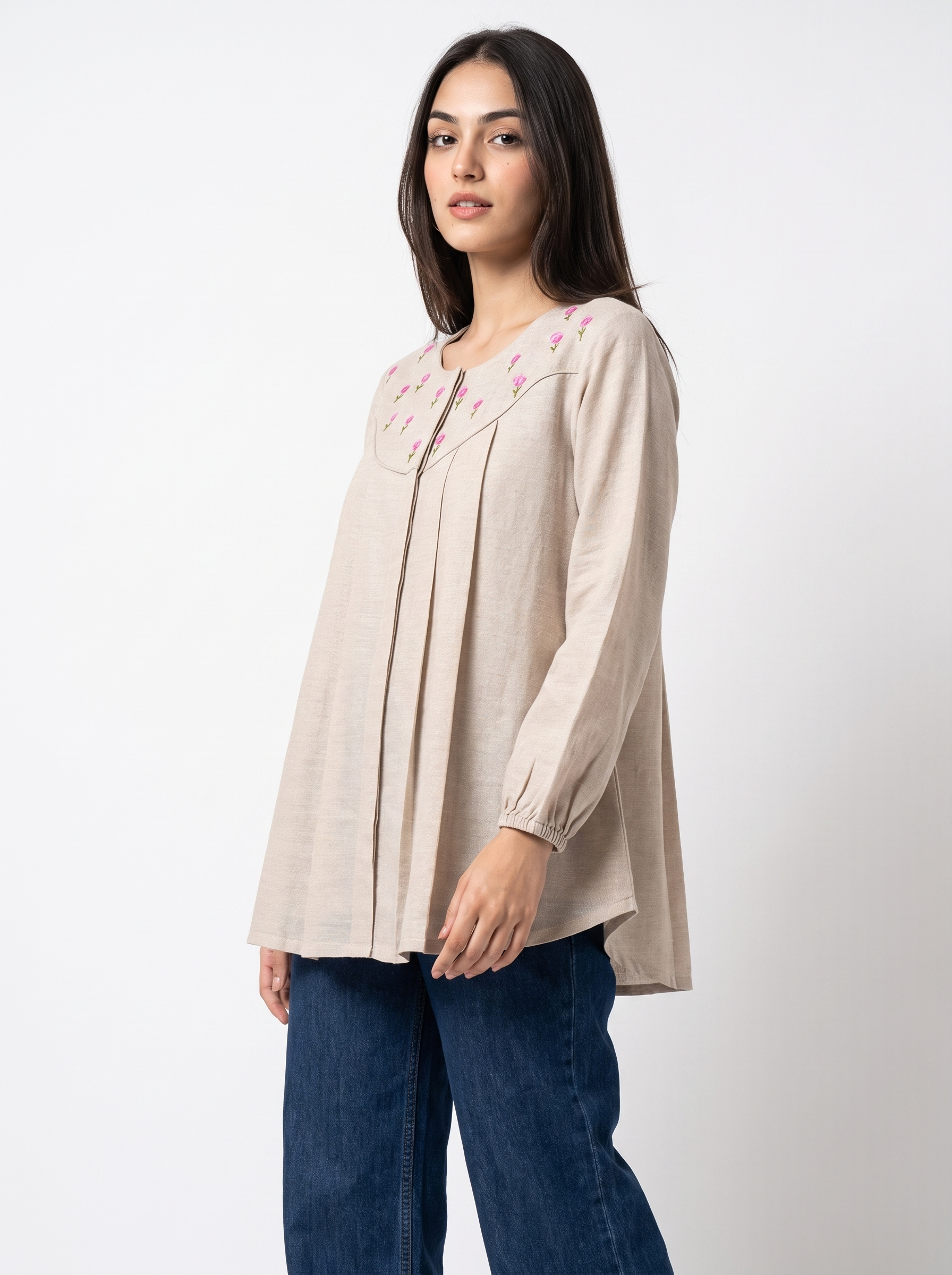 A woman modeling a beige long-sleeve tunic top with pink floral embroidery around the neckline, paired with dark blue jeans, against a plain white studio background.