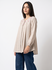 A woman modeling a beige long-sleeve tunic top with pink floral embroidery around the neckline, paired with dark blue jeans, against a plain white studio background.
