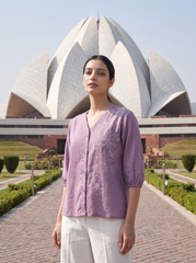 A woman stands outdoors in front of the Lotus Temple, wearing a lavender embroidered linen top and white trousers.