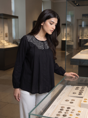 A woman in a black long-sleeve blouse with silver embroidery around the neckline and white trousers, standing in a museum next to a glass display case.