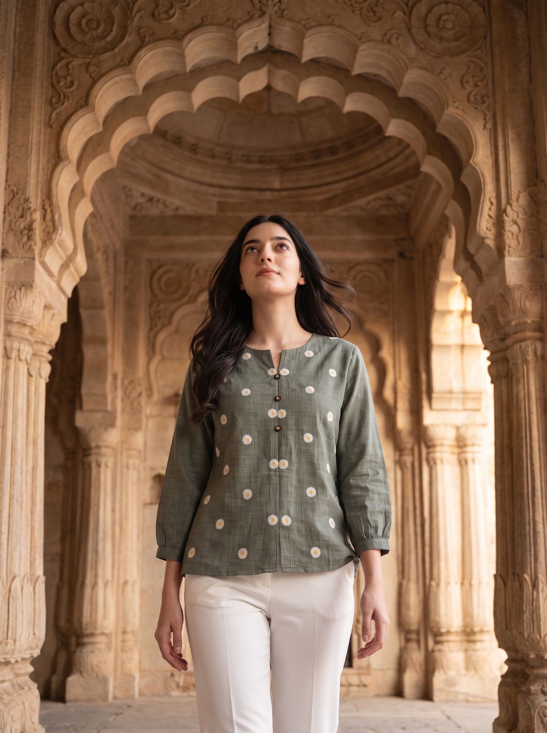 A woman wearing a sage green linen kurta with cream-colored floral prints and white trousers, standing in front of an ornate stone archway.