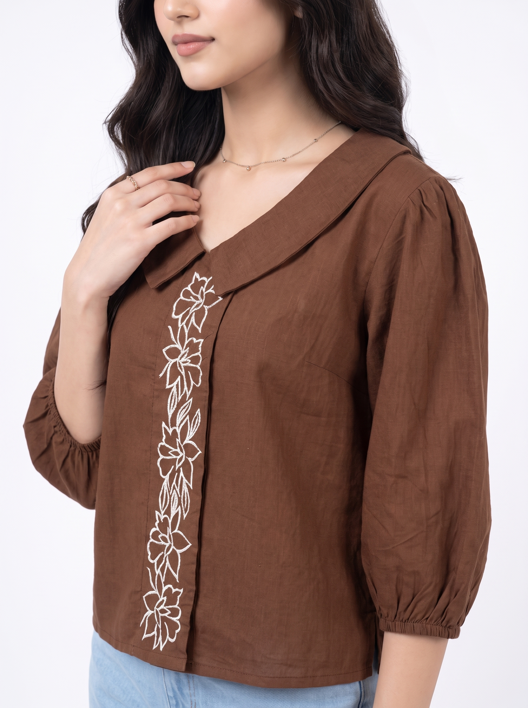 A woman wearing a brown wrap-style blouse with white floral embroidery down the front, paired with light blue jeans against a plain white background.
