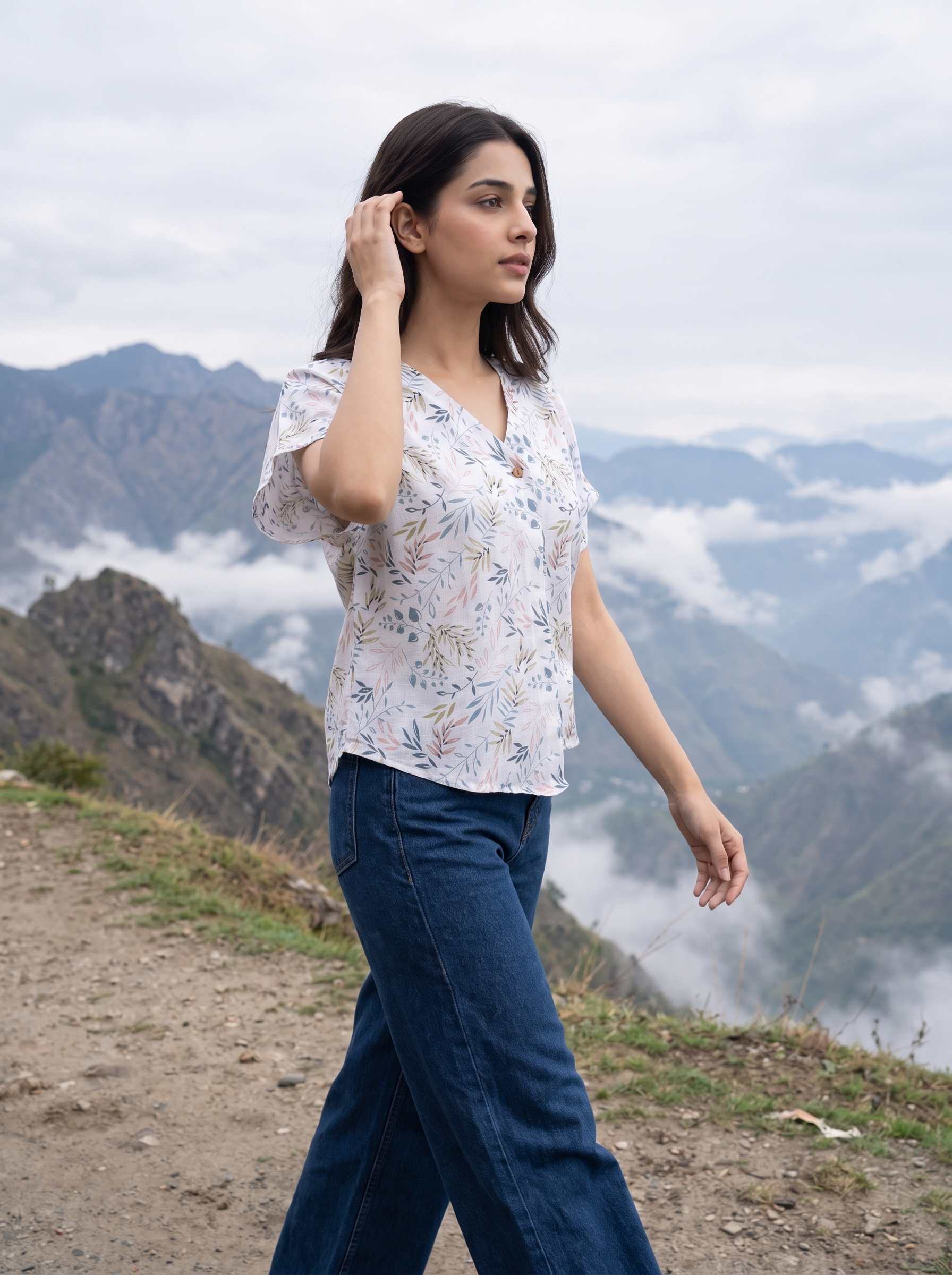 A woman wearing a white short-sleeve blouse with a delicate blue and green floral print, paired with dark blue jeans, walking along a mountain path with misty peaks in the background.