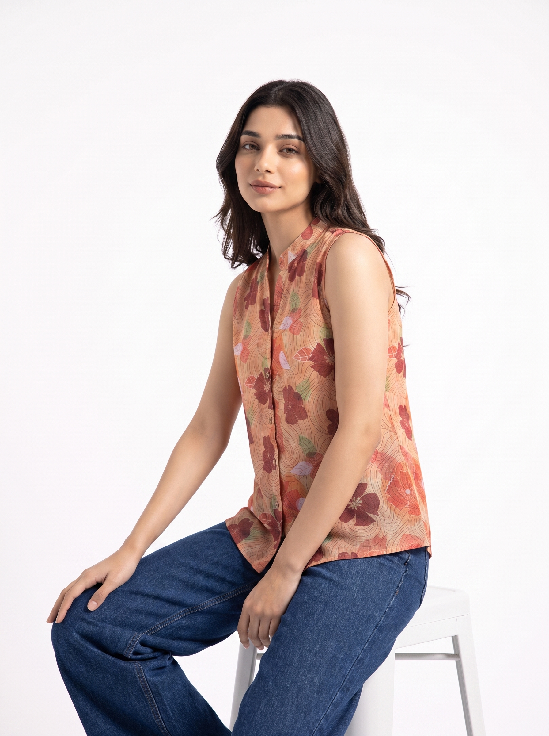 A woman wearing a sleeveless, high-neck floral top in muted orange, red, and green tones, paired with blue jeans, sitting on a white stool against a plain white background.