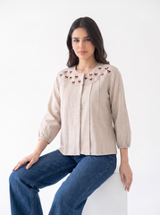 A woman wearing a beige linen blouse with red embroidered cherry details on the chest, paired with blue jeans, sitting on a white block against a white studio background.