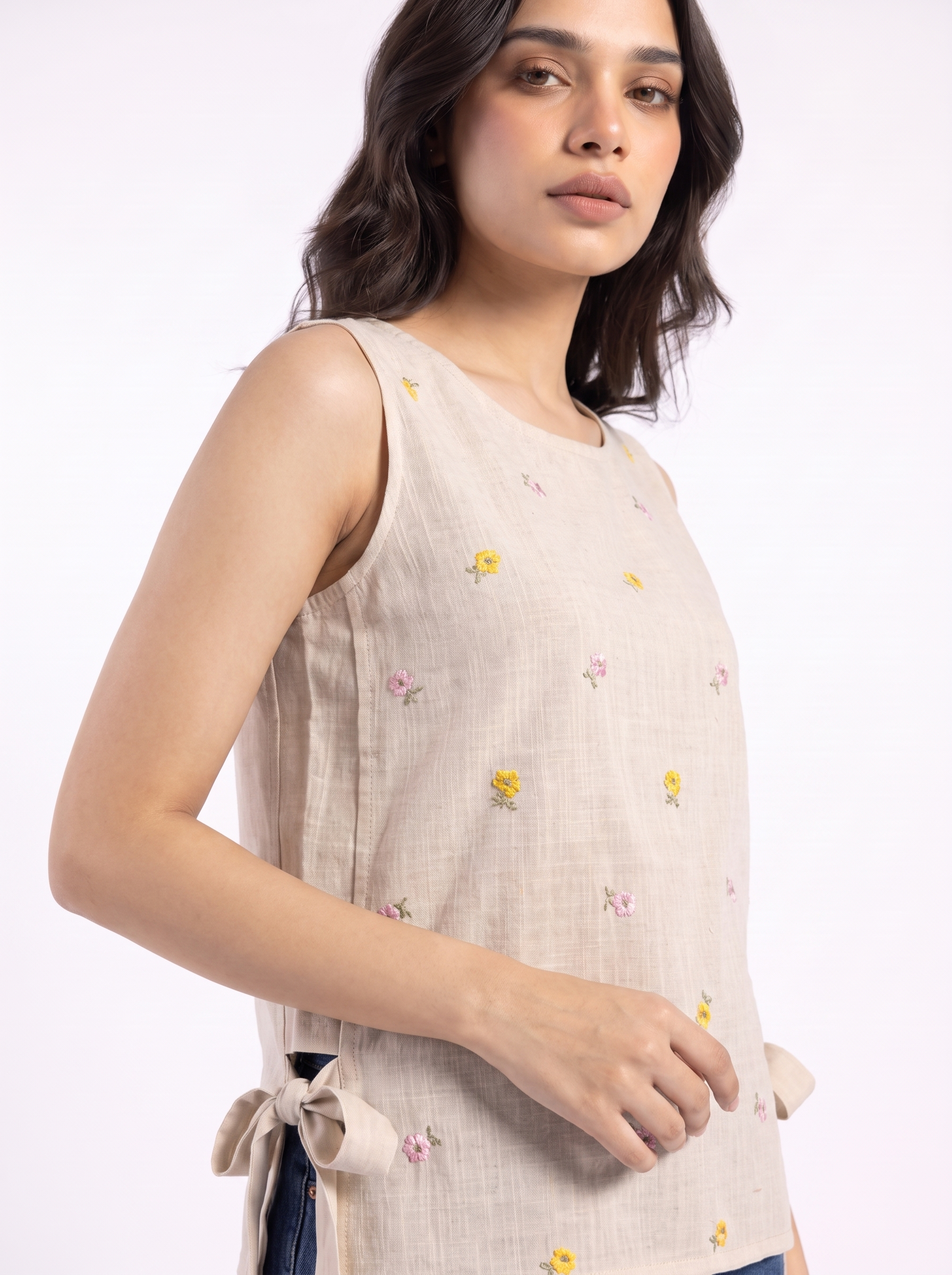 A woman wearing a sleeveless beige linen top with small embroidered yellow and pink flowers, shown against a plain white studio background.