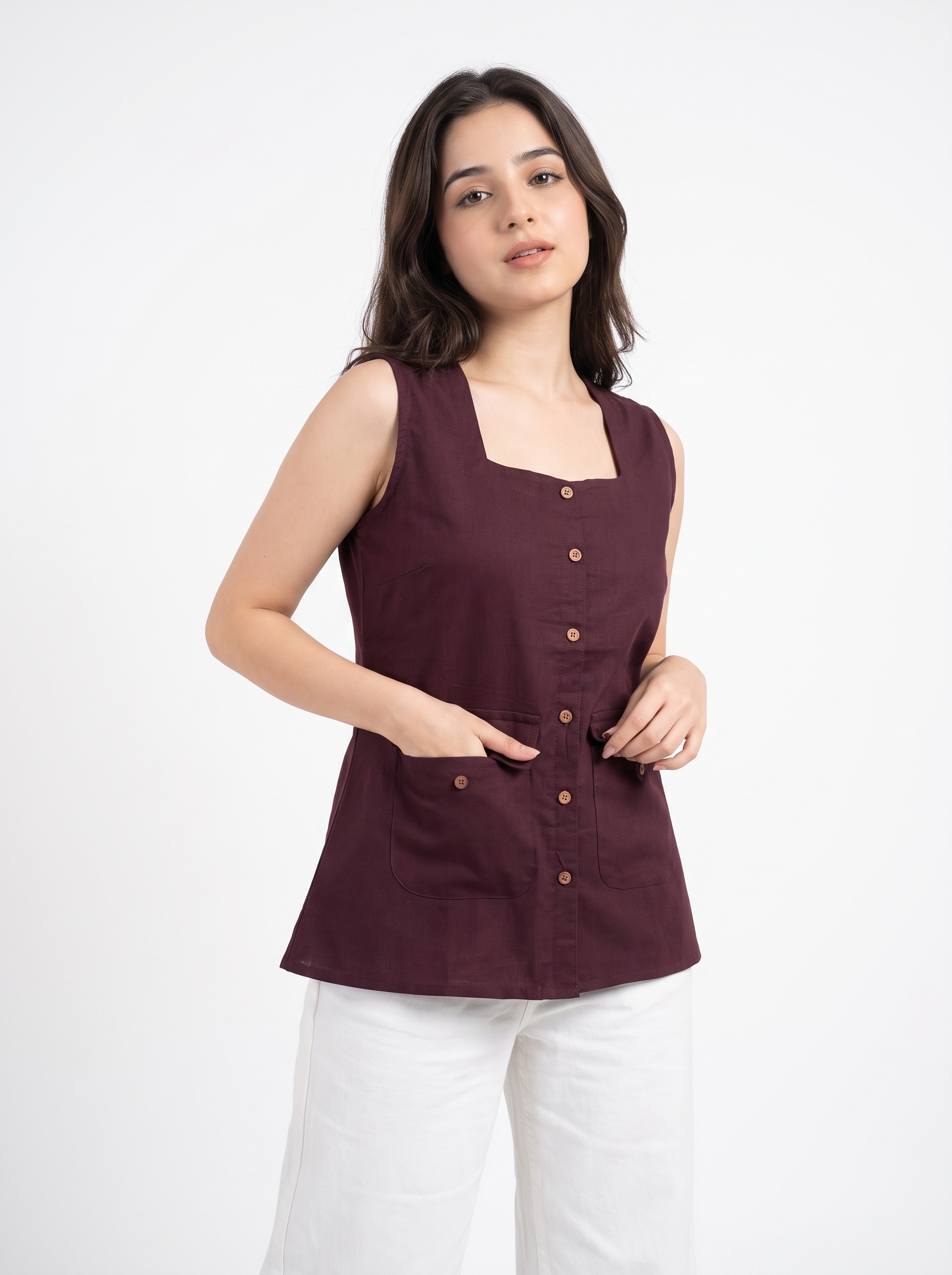 A woman modeling a sleeveless, square-neck top in deep burgundy with brown buttons, paired with white pants, against a plain white studio background.