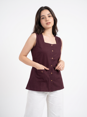 A woman modeling a sleeveless, square-neck top in deep burgundy with brown buttons, paired with white pants, against a plain white studio background.