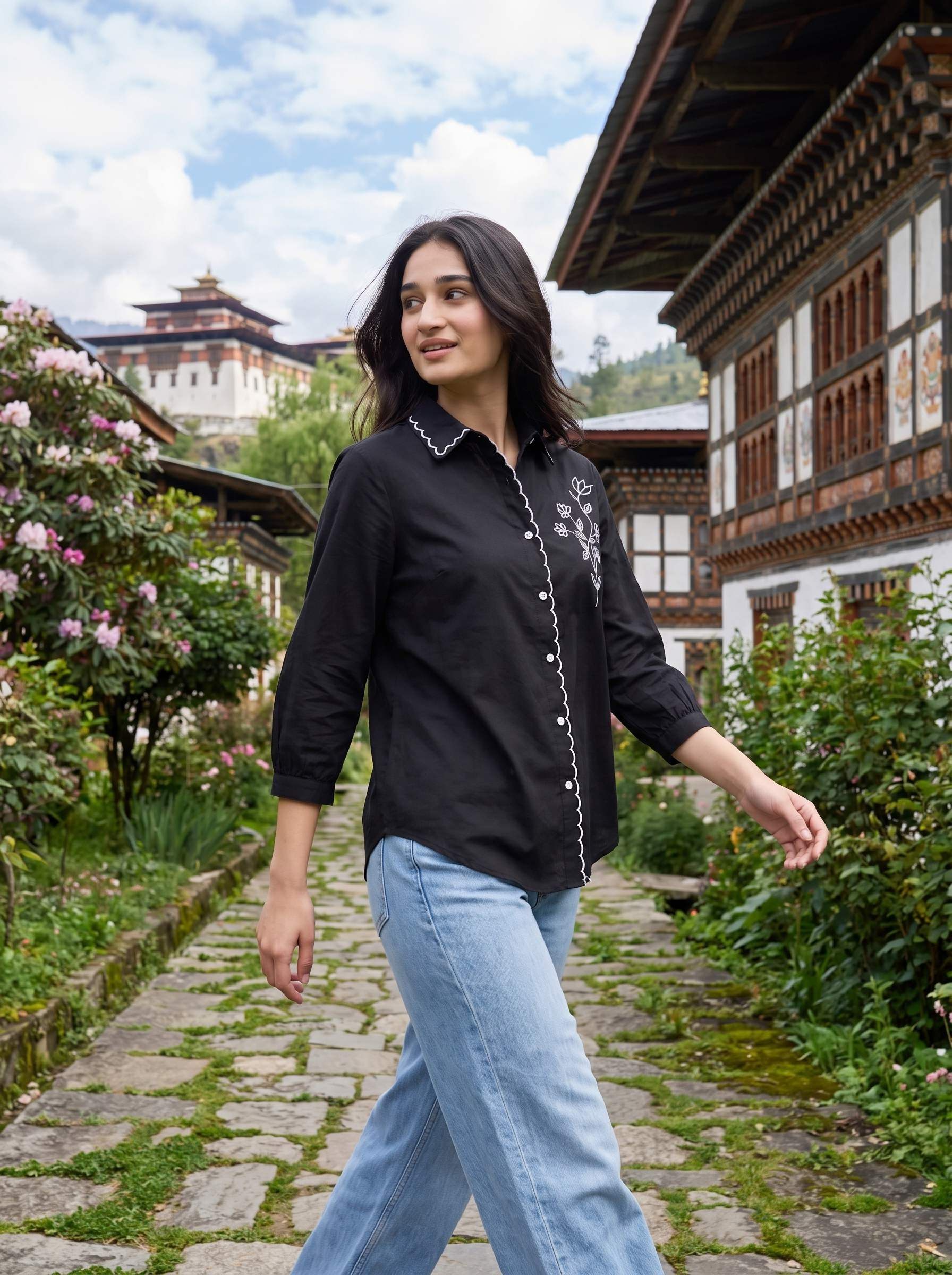 A woman wearing a black long-sleeve button-up shirt with white embroidery and light blue jeans, walking along a stone path in a traditional village setting with buildings and greenery in the background.