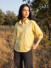 A woman wearing a pale yellow embroidered button-up shirt and dark jeans, standing outdoors in a dry grassy field with trees in the background.