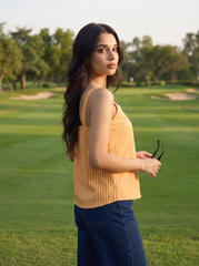 A woman standing on a golf course wearing a yellow and orange vertically striped sleeveless top and dark blue jeans, holding a pair of sunglasses.