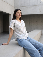 A woman wearing a white short-sleeve V-neck blouse with a delicate blue and green floral pattern, paired with light-wash blue jeans, sitting on a concrete ledge in a modern architectural setting.