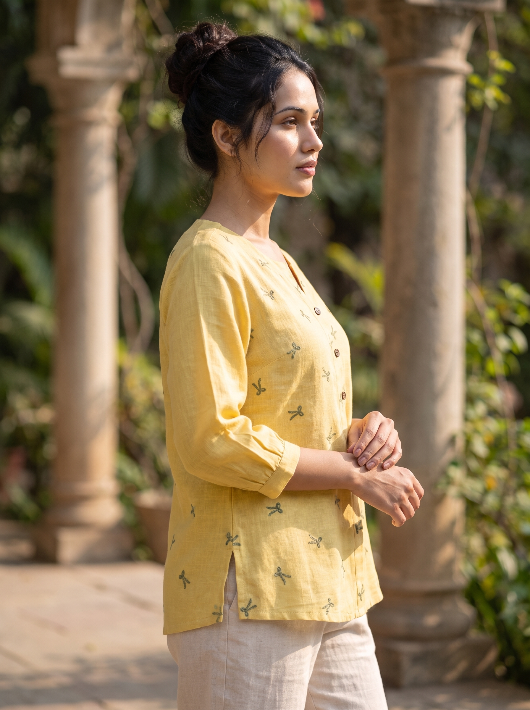 A woman wearing a light yellow linen button-up top with small green dragonfly prints and off-white trousers, standing outdoors in front of stone pillars and green foliage.