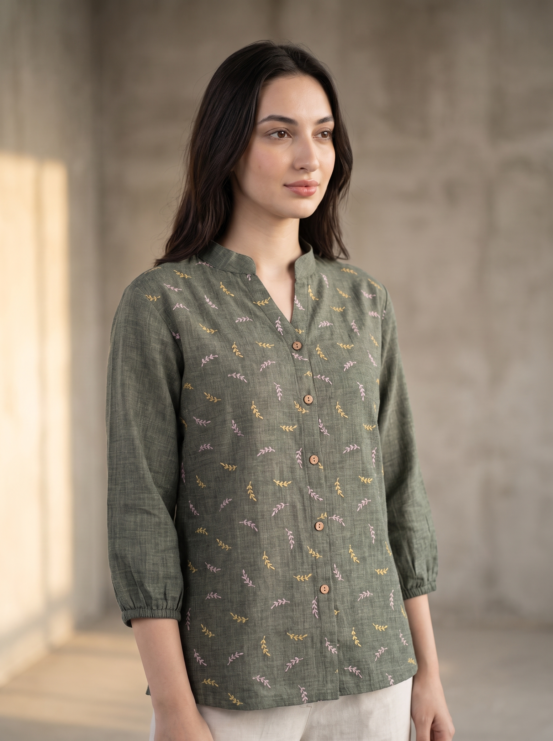 A woman wearing a sage green linen button-up shirt with a delicate multicolor floral pattern, standing in a minimalist concrete room.